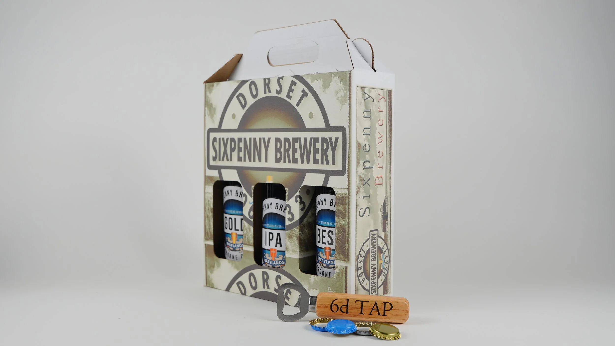 Sixpenny Brewery beer bottles in a cardboard carrier, labeled Gold, IPA, and Bes, with a wooden beer tap labeled "6d TAP" and some bottle caps in front.