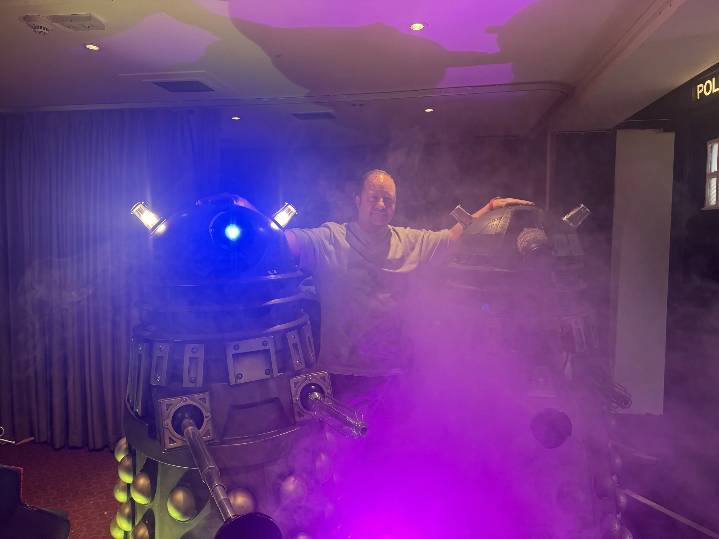 A man standing between two life-sized Dalek robots from Doctor Who, with purple and blue lighting and fog.