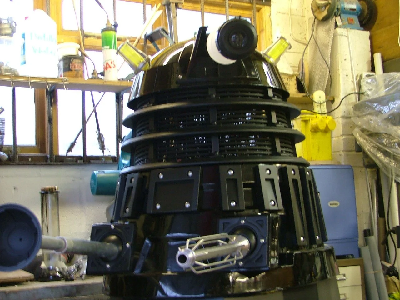A detailed model of the Dalek, a fictional extraterrestrial race from Doctor Who, is displayed in a workshop. It has a black lower body with silver accents, a dome-shaped head with a light, and a protruding arm and gunstick at the front.