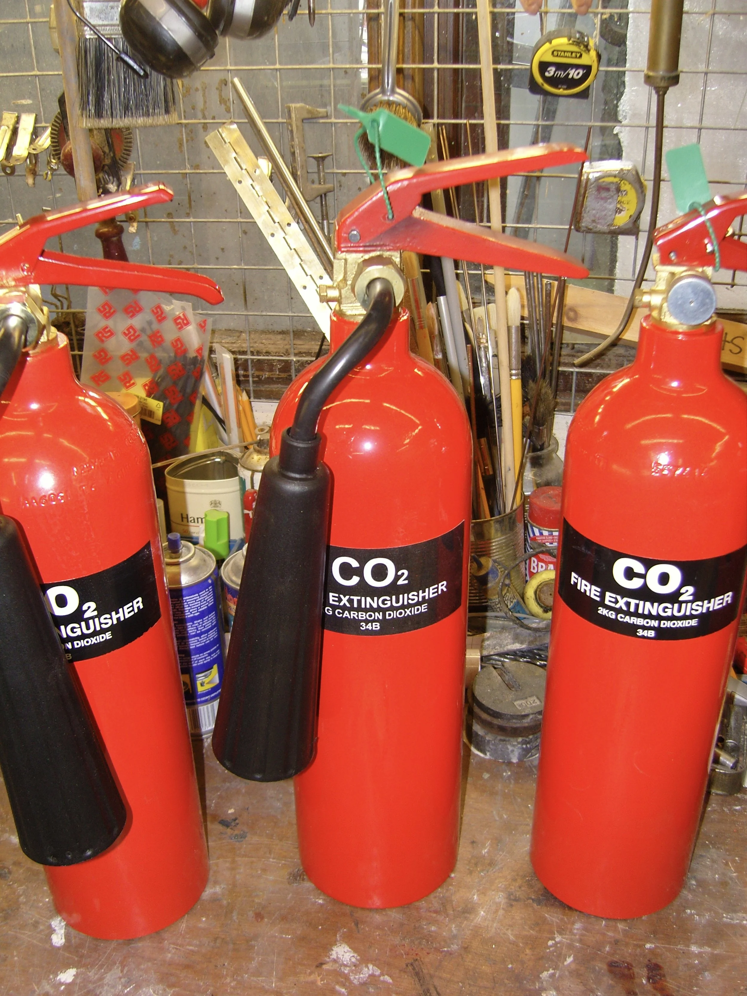 Three CO₂ fire extinguishers on a cluttered workshop workbench with tools and supplies in the background.