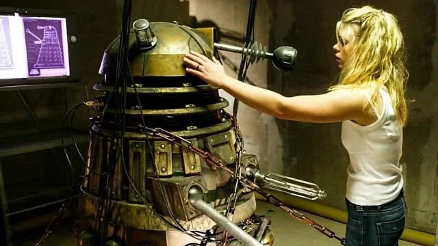 A woman with blonde hair wearing a white shirt and jeans is interacting with a Dalek, a fictional extraterrestrial mutant from Doctor Who, in a science fiction setting.