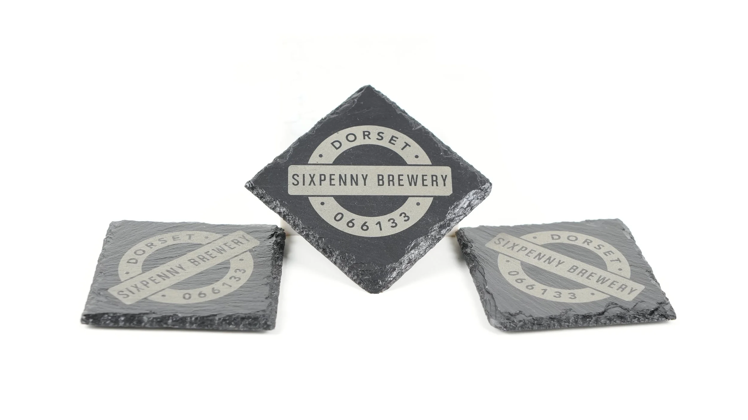 Three black slate coasters with circular and rectangular printed labels showing branding for Sixpenny Brewery, Dorset, and the phone number 066133. One coaster is standing upright, the other two are lying flat.