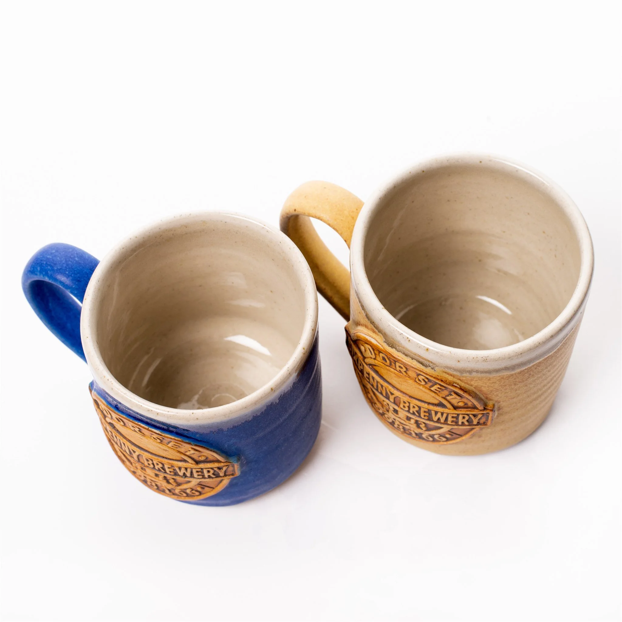 Two ceramic beer steins with brewery logos, one blue and one beige, viewed from above on a white background.
