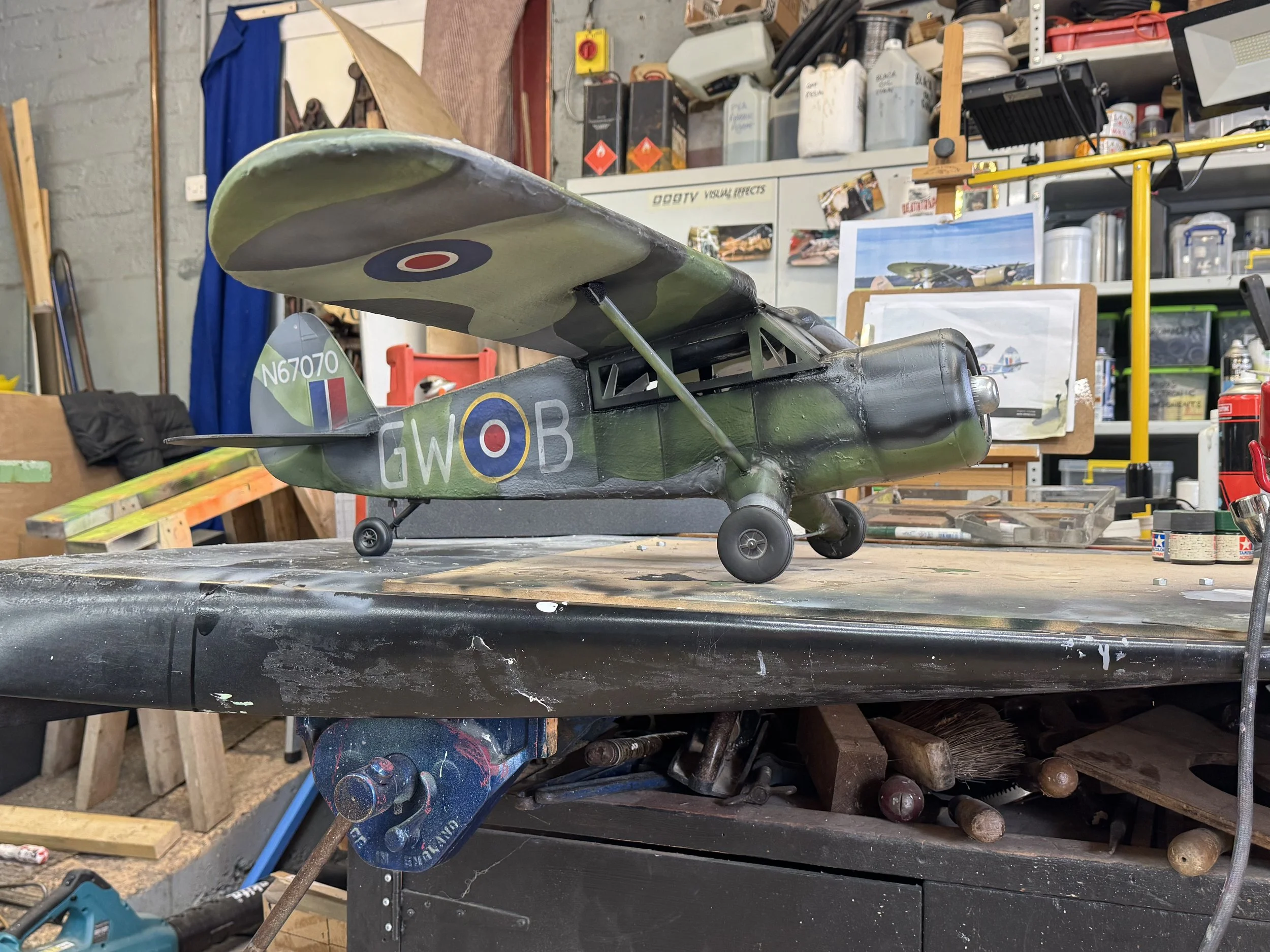 Model of a vintage military helicopter with camouflage paint, displayed on a workbench in a workshop surrounded by tools and supplies.