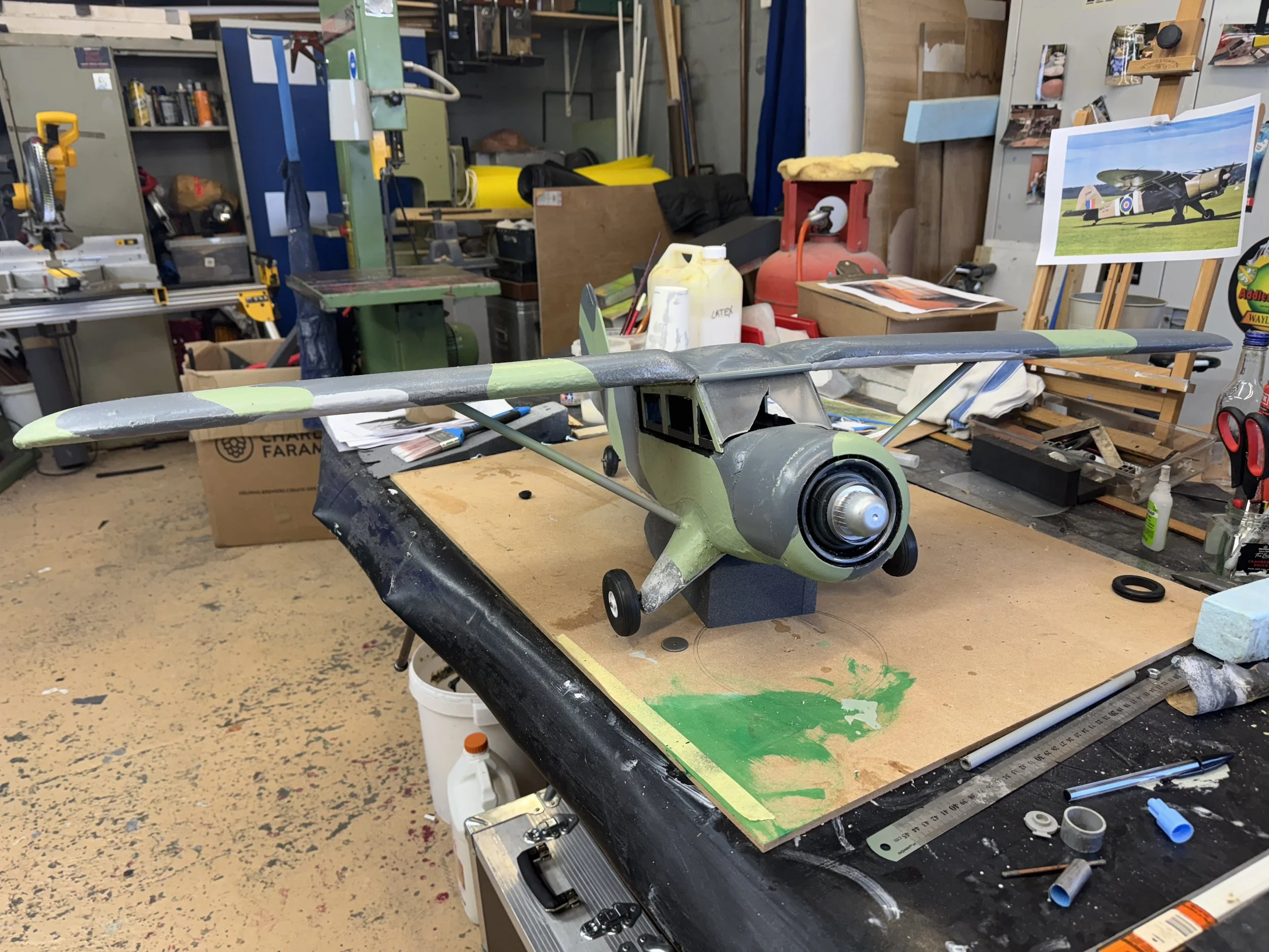 A detailed scale model of a vintage aircraft on a worktable in a workshop. The model has a green and gray color scheme, with a large front engine and wings extending on either side. The workshop contains tools, paint, photos of vehicles, and various 