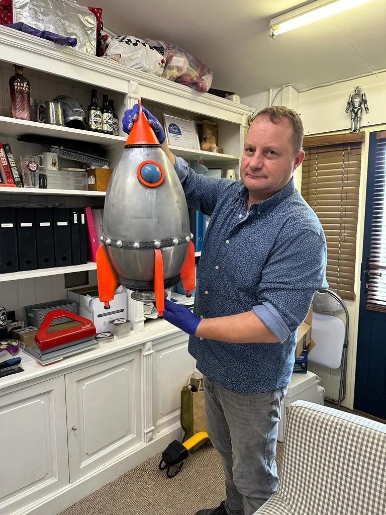 A man stands indoors holding a model rocket designed to resemble a cartoonish space rocket with a cone-shaped top, metallic body, orange fins and details, and a blue nose. The man wears a blue shirt, gray pants, and blue gloves.