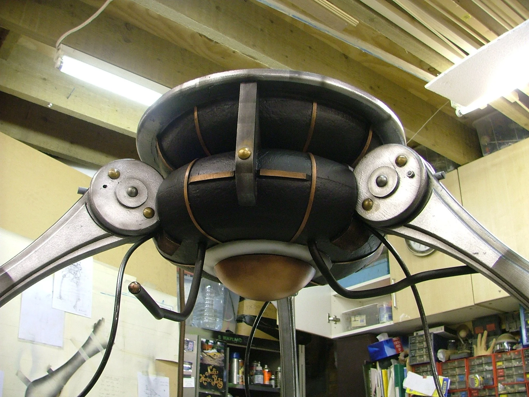 Close-up view of a metallic robot head with mechanical joints and wires, set indoors with shelves and a wall clock in the background.