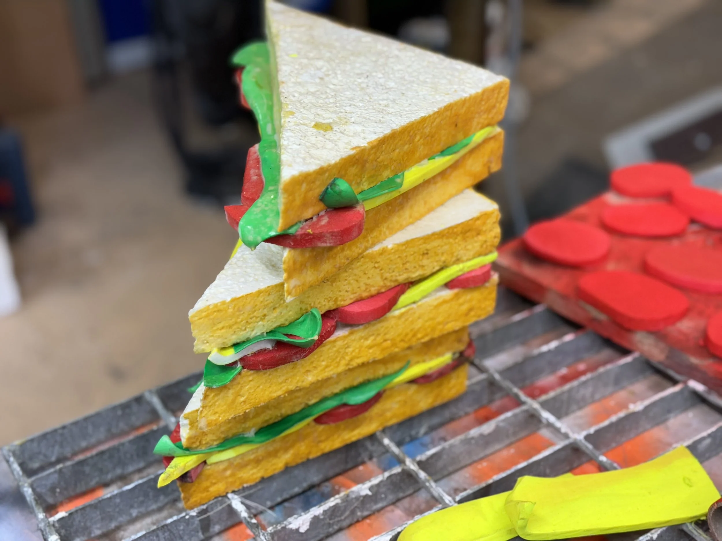 Colorful craft sandwich made of foam or cardboard with yellow bread, red and green toppings, assembled in a stacked design on a metallic surface, with red foam hearts on a tray in the background.