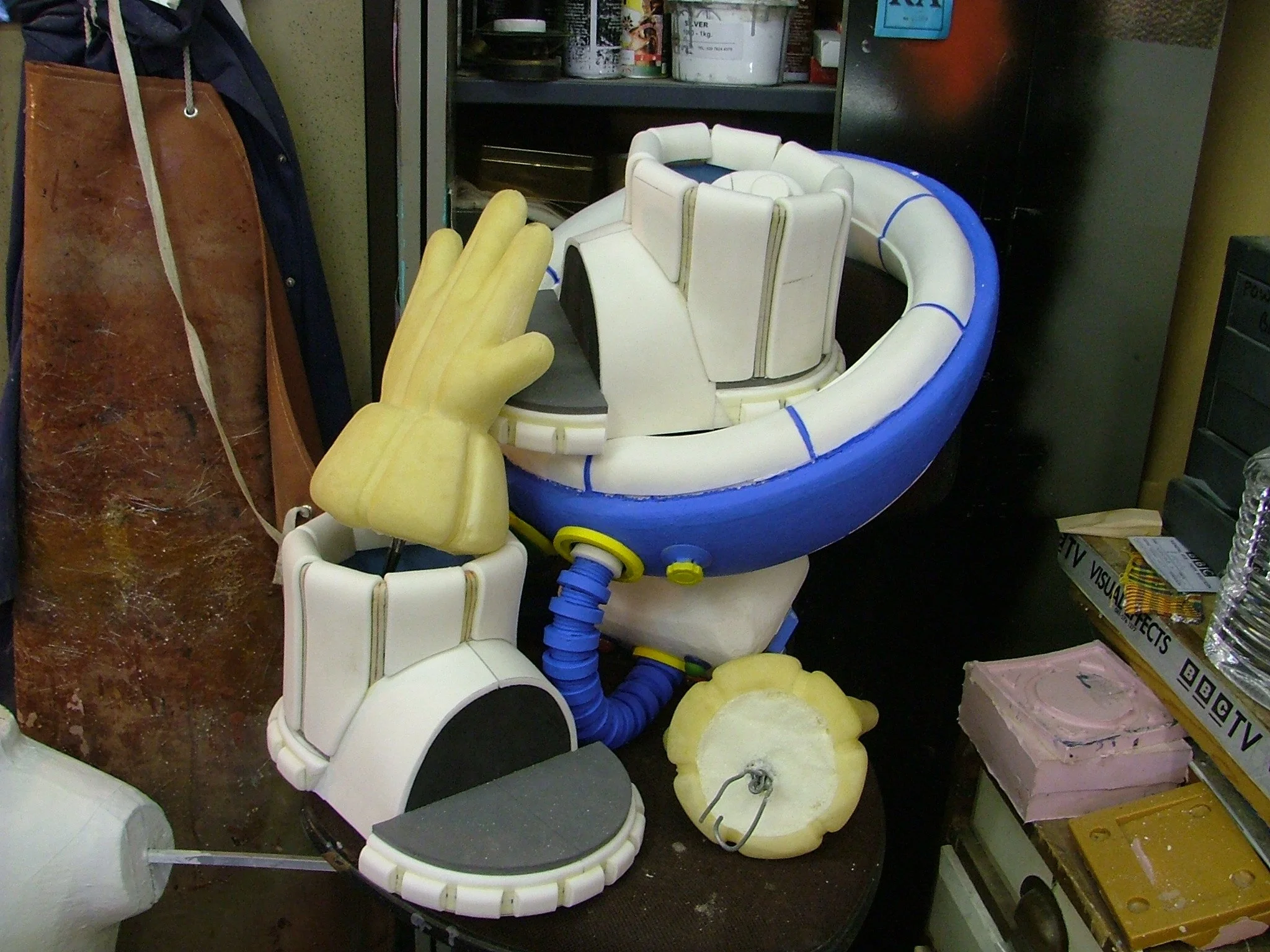 Large cosplay or costume prop resembling a robot or cartoon astronaut Lunar Jimcharacter's arm and hand, with a yellow glove on the hand, sitting on a table. Background includes various supplies and a brown corkboard.