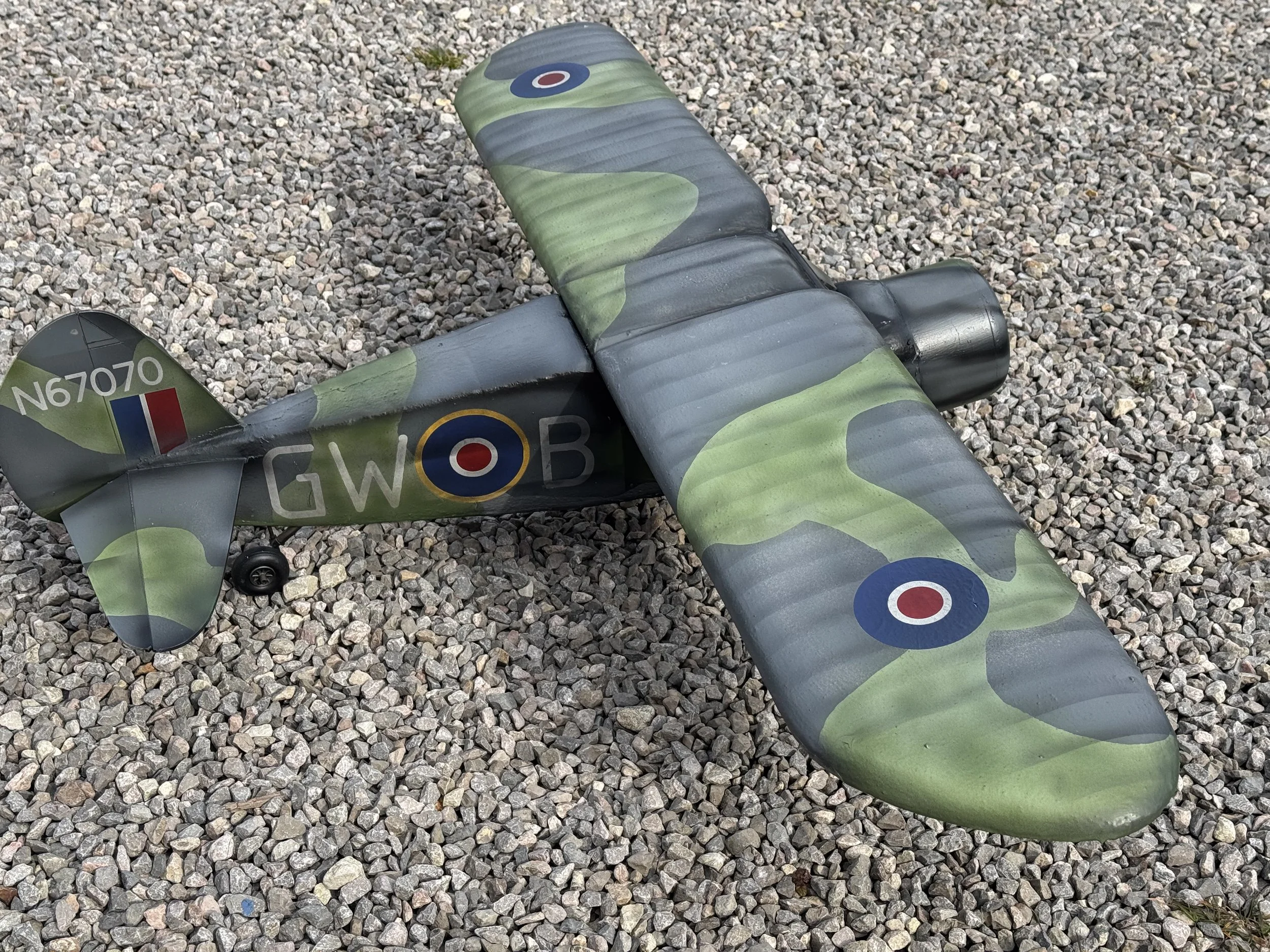A model airplane painted in a green, gray, and black camouflage pattern with Royal Air Force roundels and markings, lying on gravel ground.