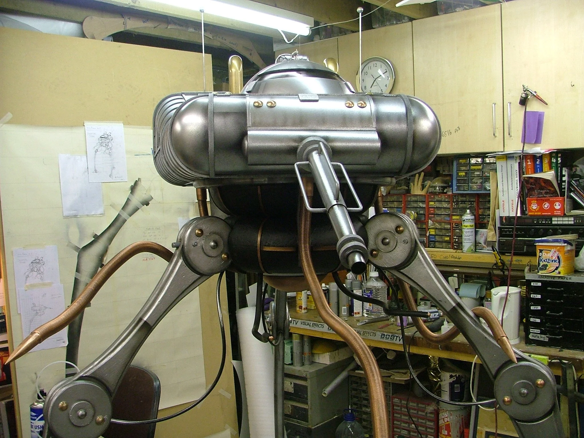 A large construction robot in a workshop with shelves and tools in the background.