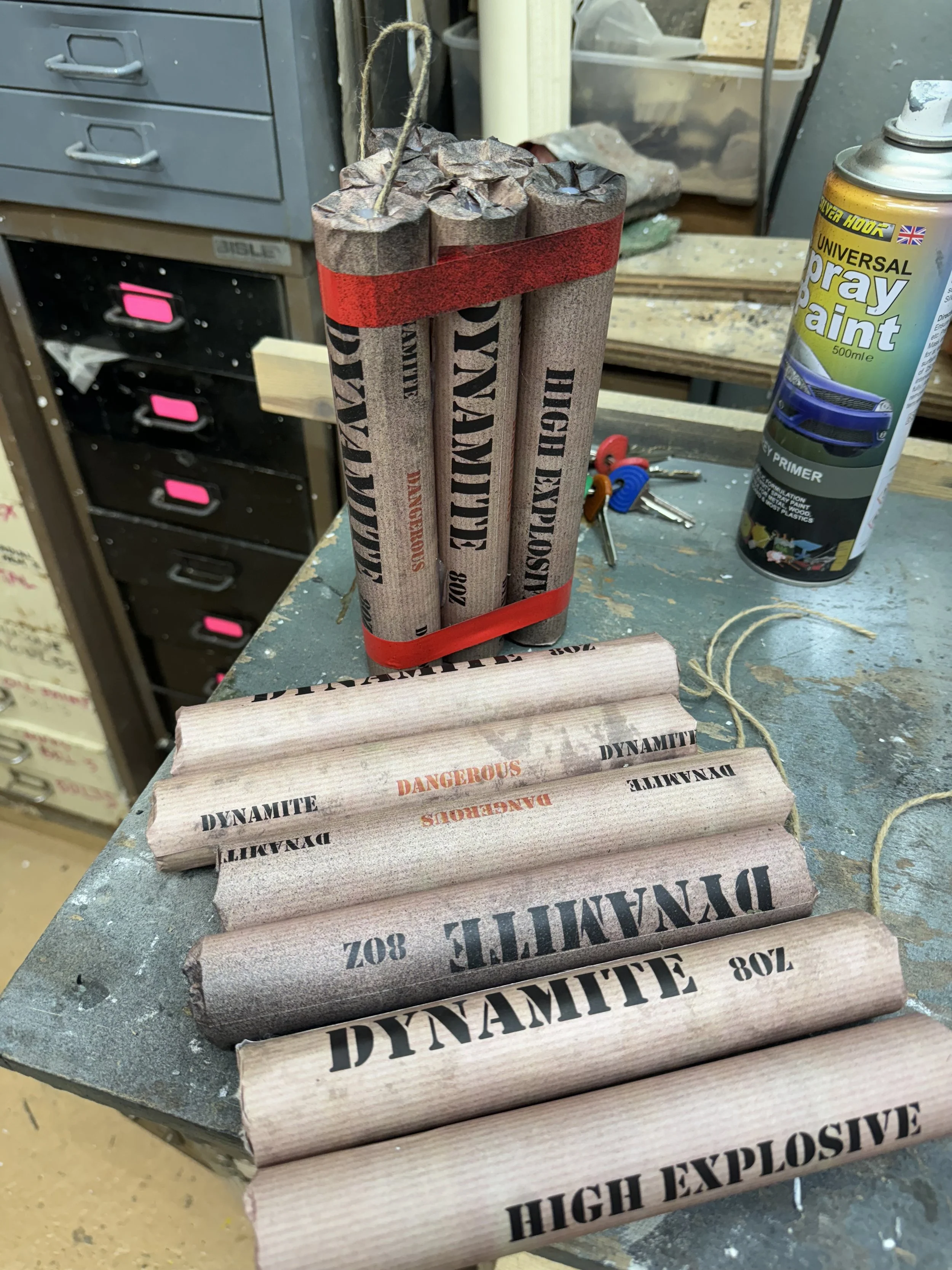 Bundles of fake prop sticks of 'DYNAMITE' and 'HIGH EXPLOSIVE' on a workbench in a workshop or garage.