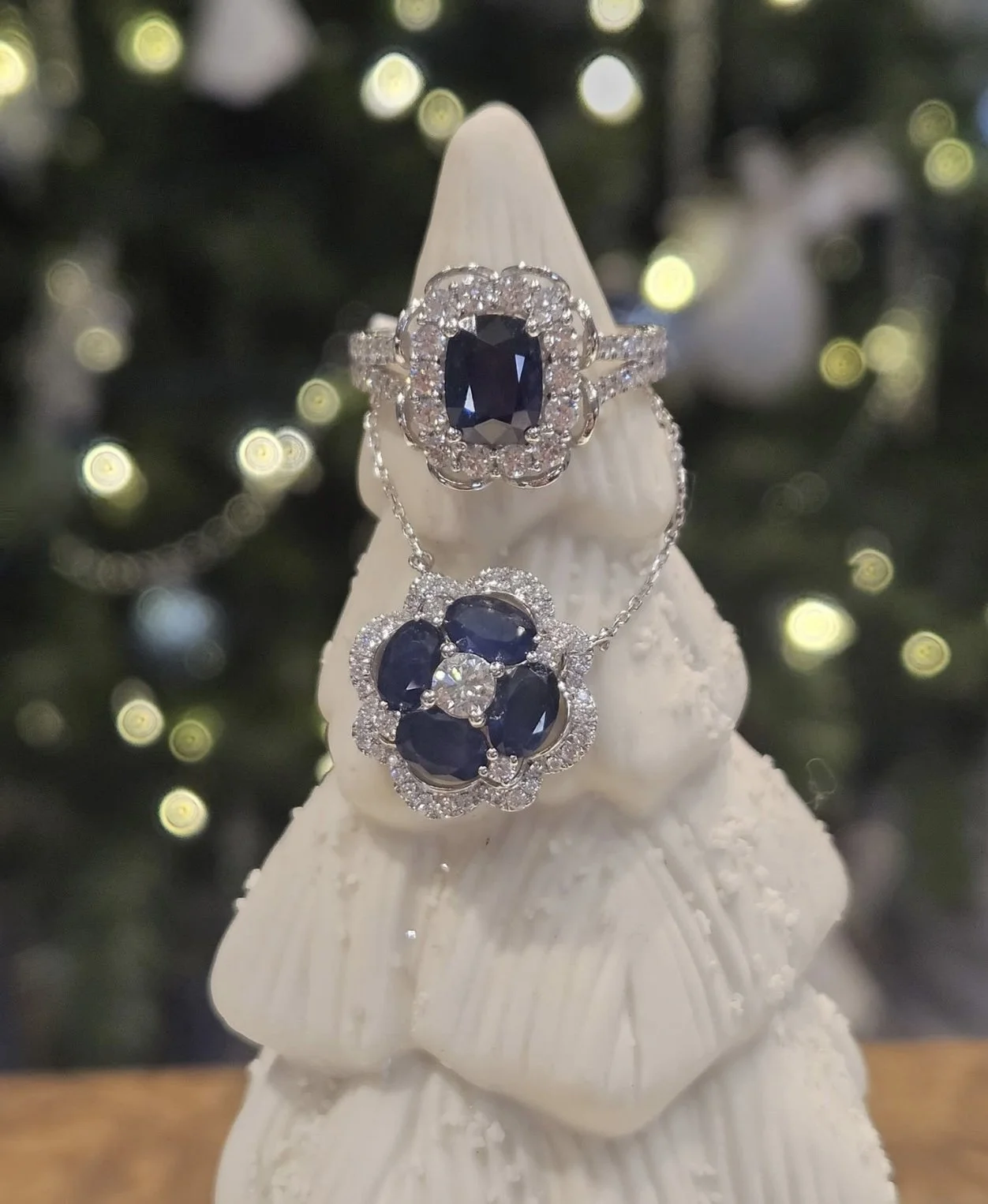 A jewelry set with a ring, necklace, and earrings featuring dark blue gemstones surrounded by small diamonds, displayed on a white Christmas tree ornament with blurred Christmas lights in the background.