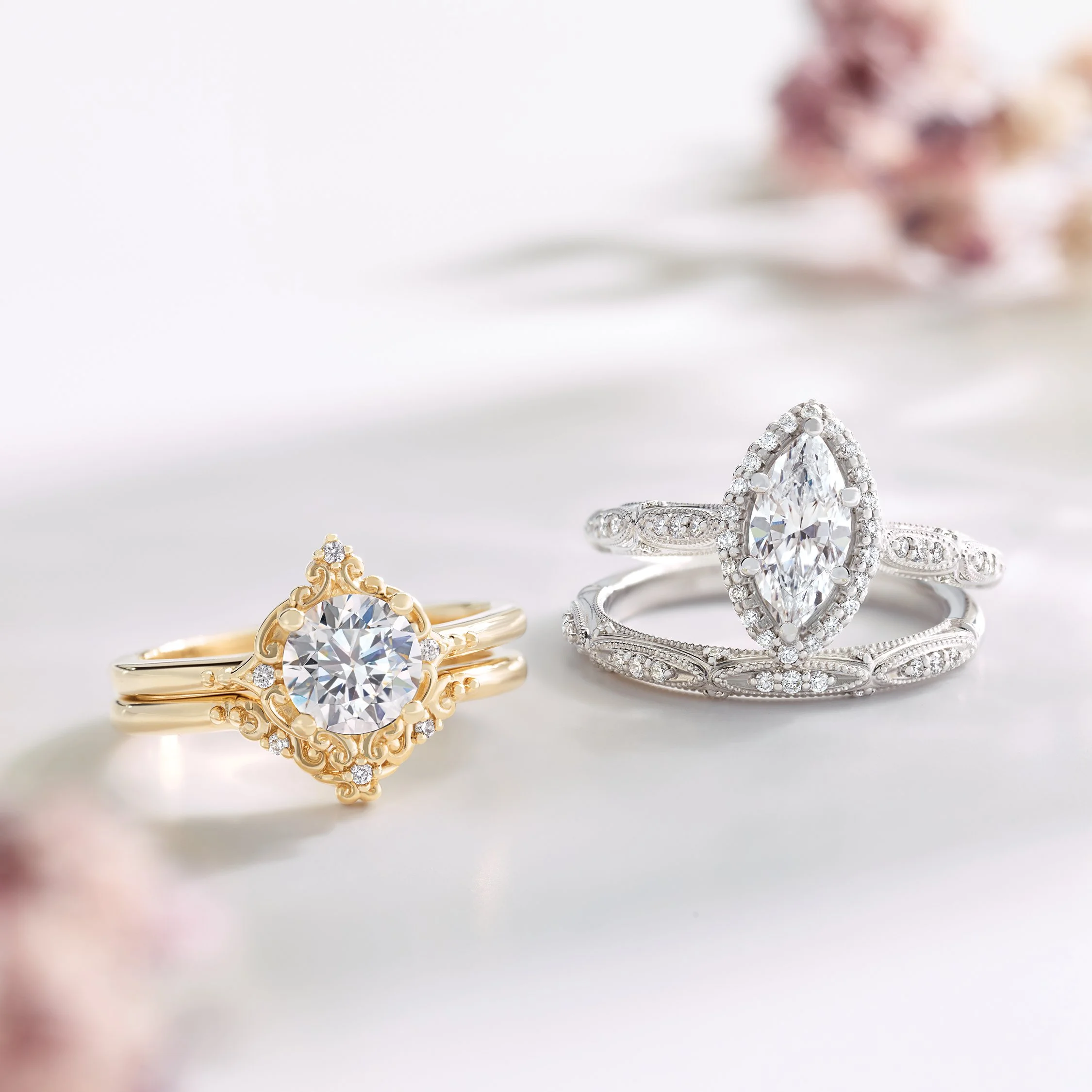 Two elegant engagement rings, one gold with intricate detailing and a large center diamond, and the other silver with a marquise-shaped diamond, both adorned with smaller diamonds, placed on a soft white surface with blurred pink flowers in the background.