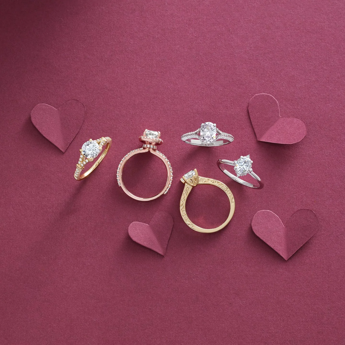 Six rings of various designs and metals arranged on a pink background with pink paper hearts scattered around.
