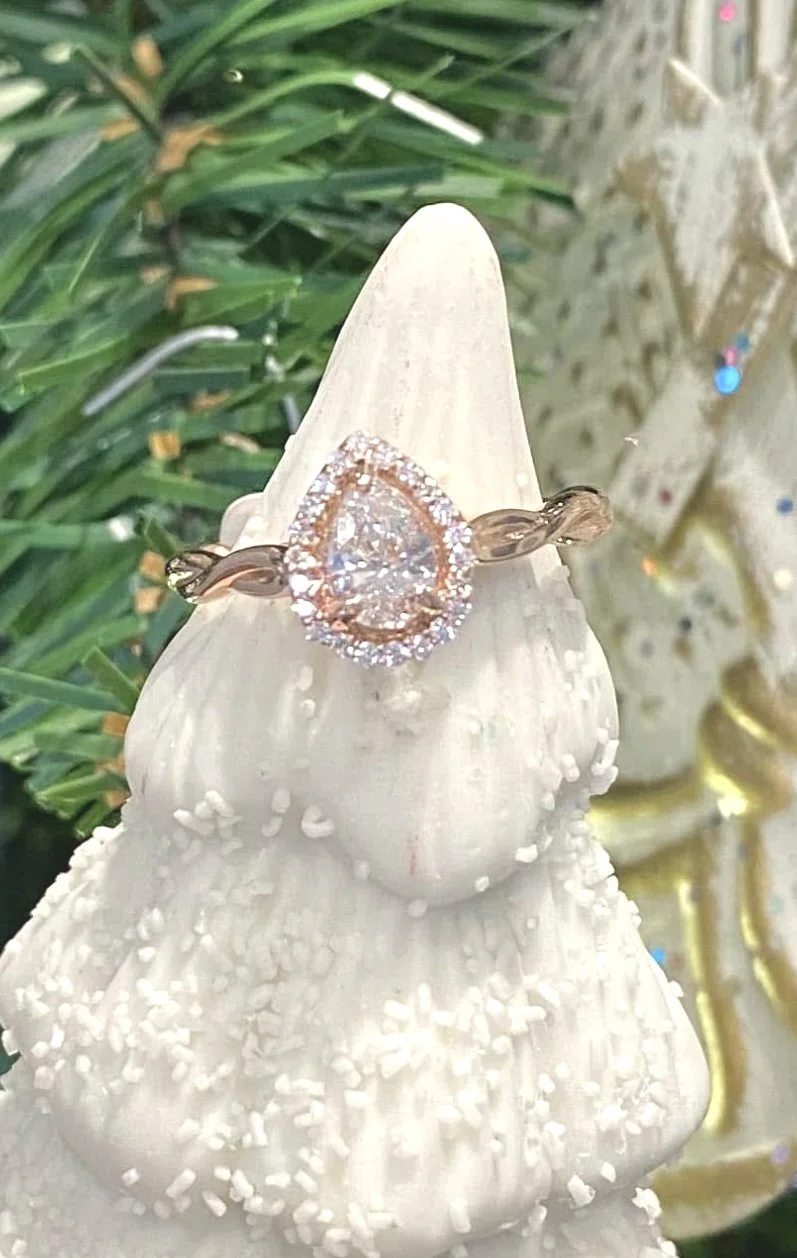 Close-up of a diamond ring with a pear-shaped center stone surrounded by smaller diamonds, displayed on a white decorative cone-shaped object with textured surface, in front of green pine branches.