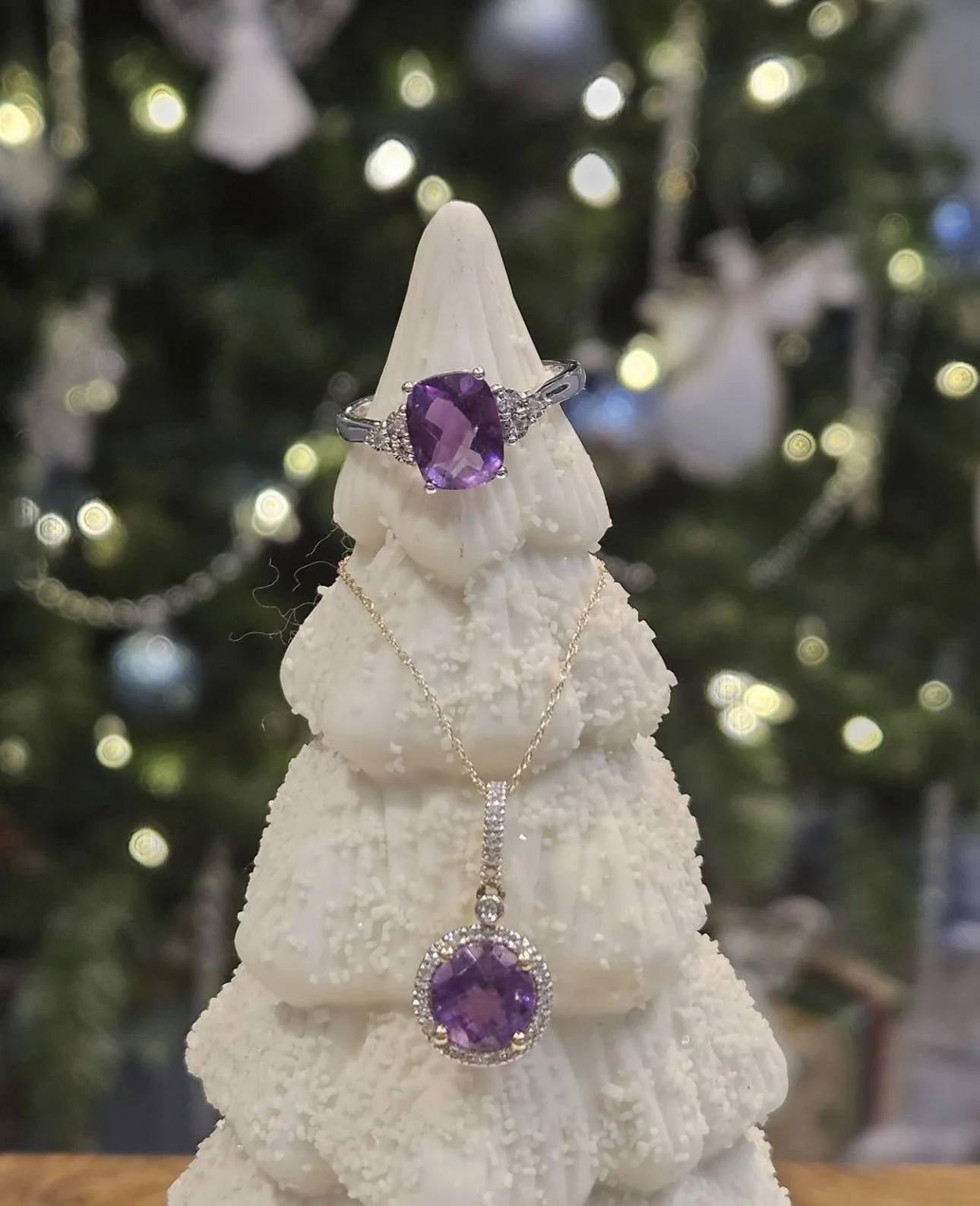 A white decorative Christmas tree with a ring and a necklace, all featuring large purple gemstones, set against a blurred background of Christmas lights.