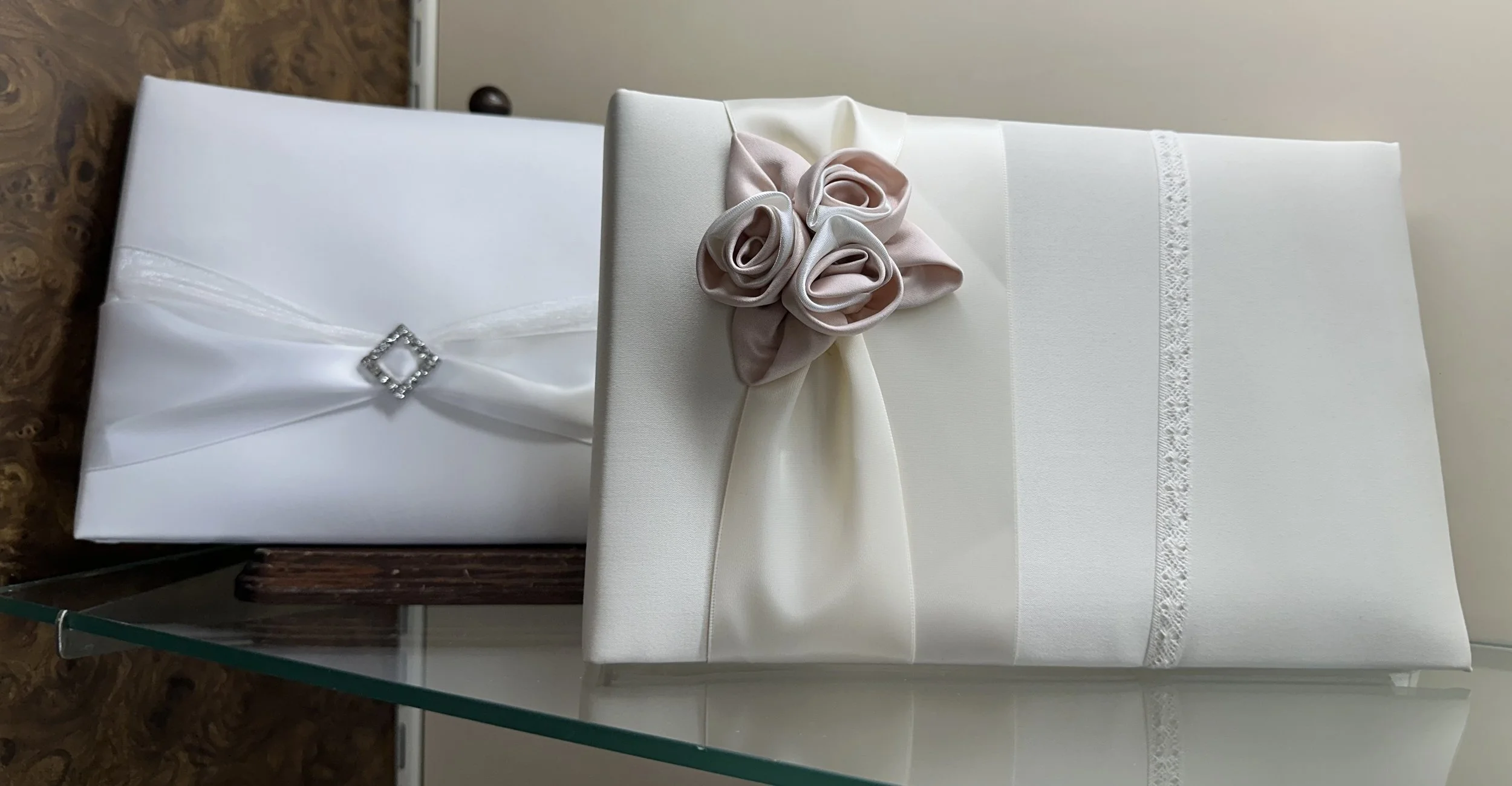 Two white gift boxes placed on a glass surface, one with a satin ribbon decorated with a rhinestone square, and the other with a ribbon and fabric roses.