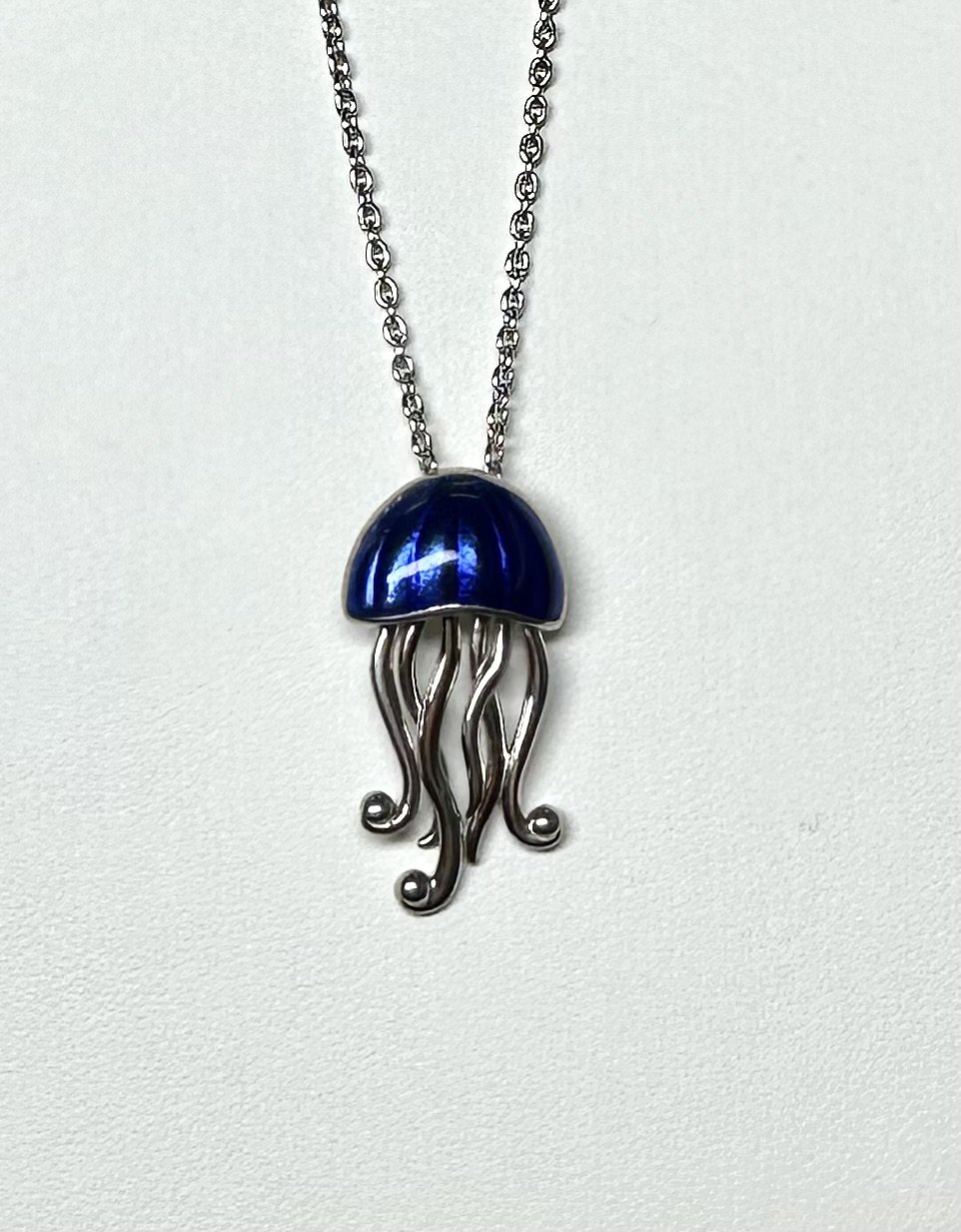 A silver necklace with a pendant shaped like a jellyfish. The jellyfish has a blue, enameled, dome-shaped top and silver tentacles extending downward.