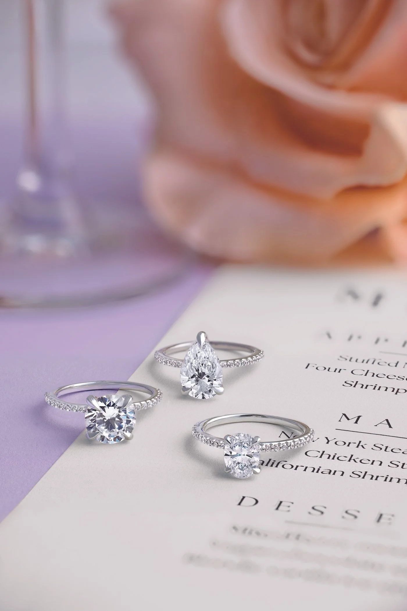 Three diamond engagement rings displayed on a piece of paper, with a soft pink flower and a blurred glass in the background.