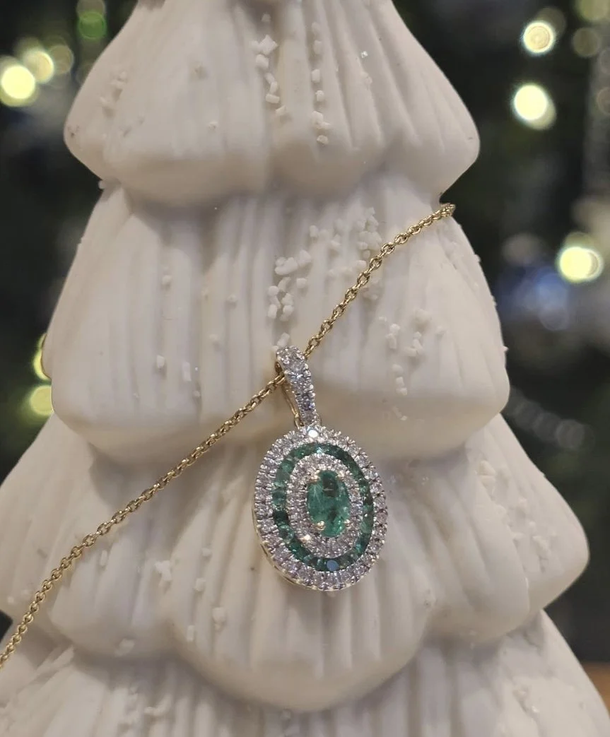 Close-up of a pendant necklace featuring a large oval-shaped green gemstone surrounded by smaller clear stones, hanging on a thin gold chain, displayed on a white ceramic figurine with textured surface, with blurred lights in the background.
