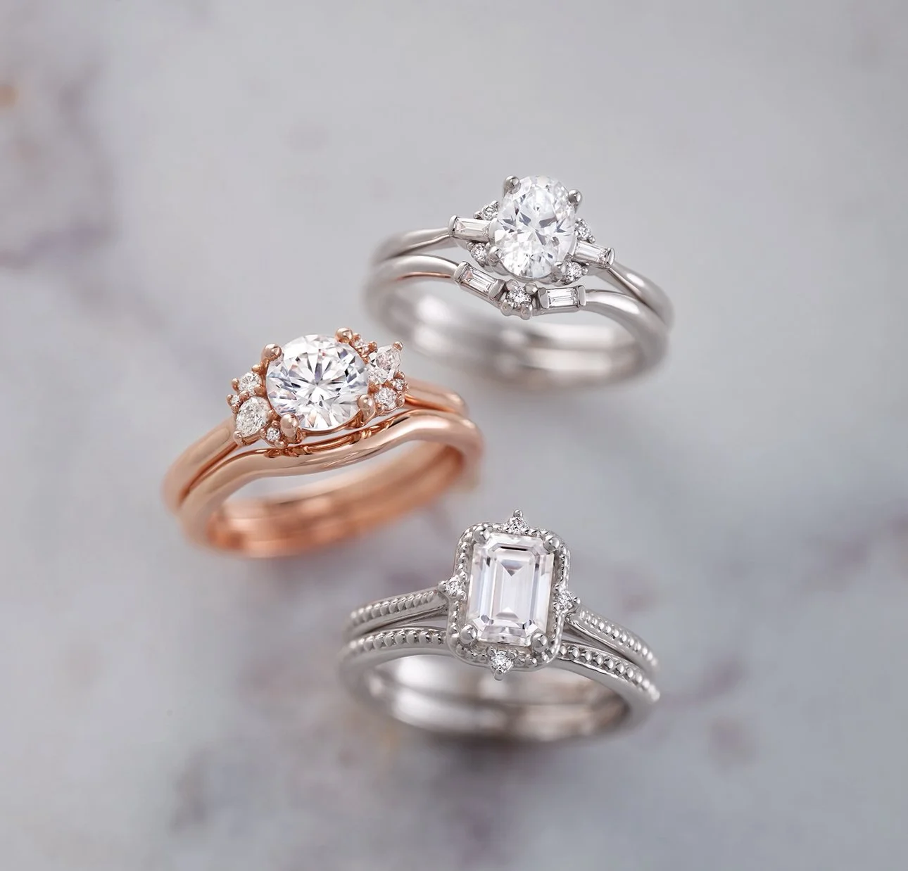 Three elegant rings, one rose gold with a large center diamond and surrounding smaller diamonds, and two silver rings with prominent center diamonds surrounded by smaller diamonds, displayed on a soft neutral background.
