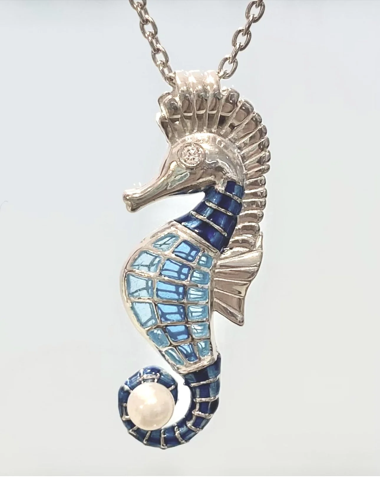 A silver pendant shaped like a seahorse with blue enamel accents and a pearl at the tail, hanging from a chain.