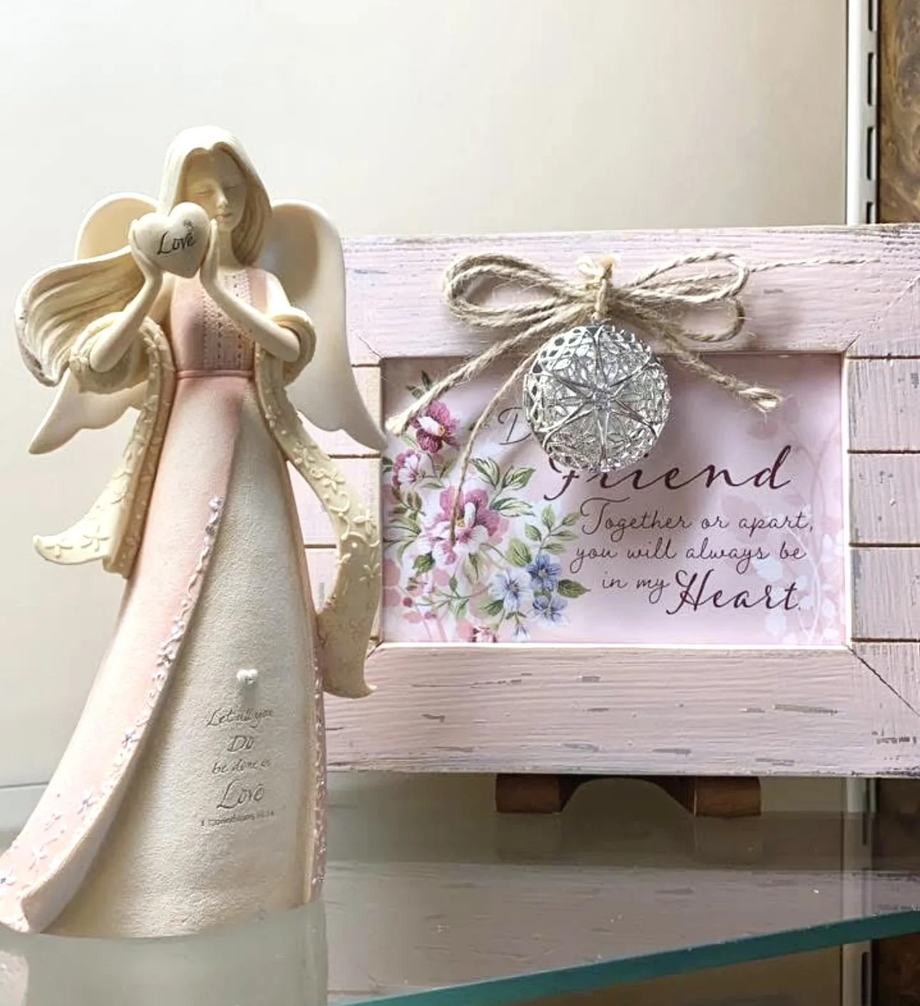 Decorative angel figurine holding a heart with the word 'Love', next to a pink wooden photo frame with a floral message and a silver ornament tied with a twine bow.