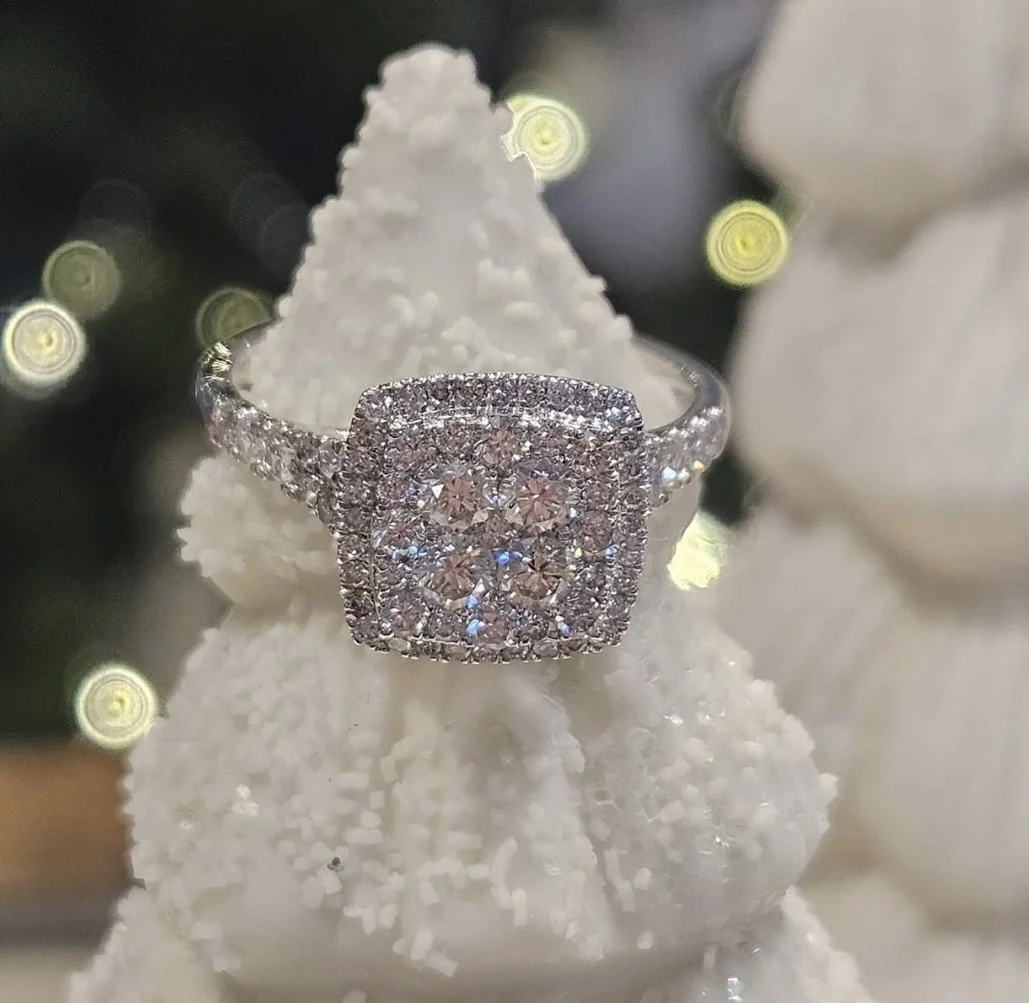 A silver ring with multiple small diamonds displayed on a snowy white Christmas tree ornament with blurred lights in the background.