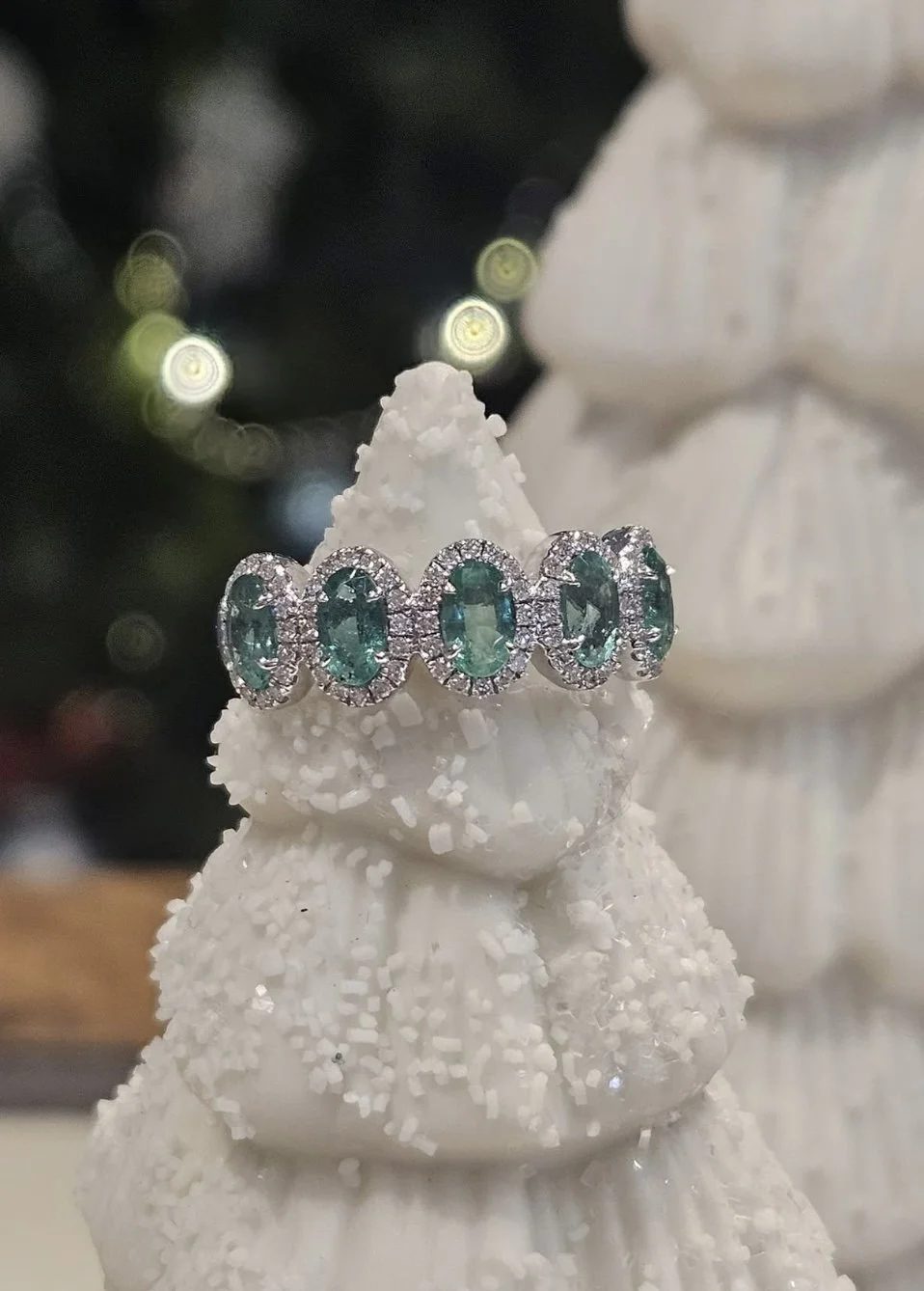 A silver ring with oval emerald gemstones and small diamonds adorns the top of a decorated white Christmas tree.