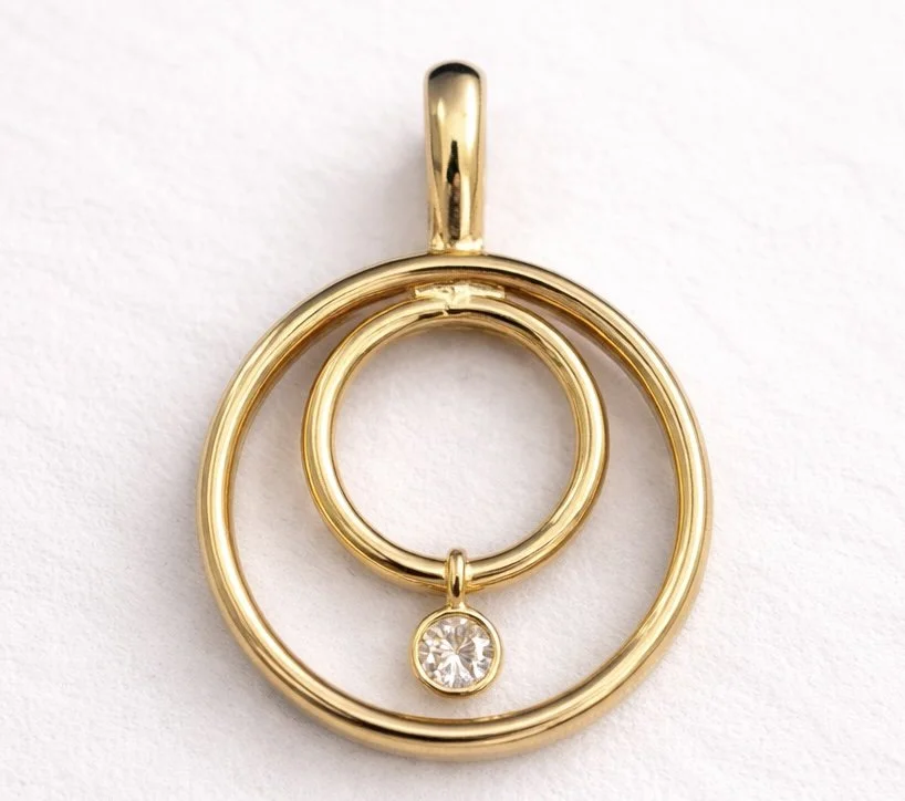 Gold pendant necklace with three concentric circles, one small inner circle, and a dangling diamond-shaped gemstone.