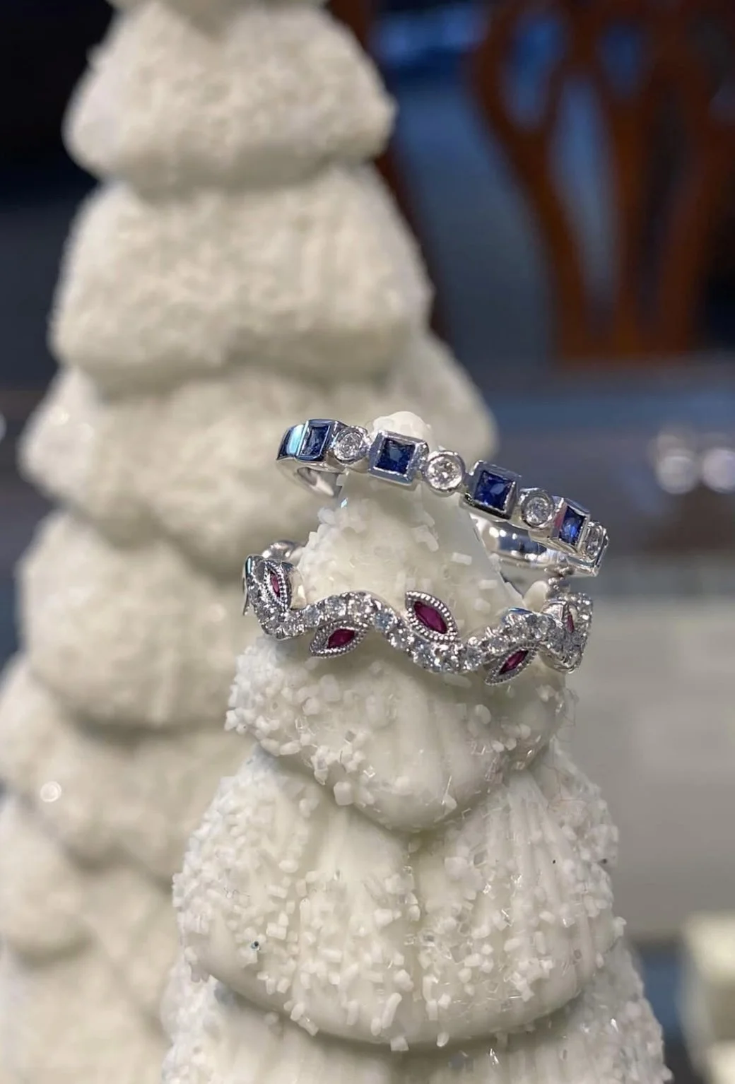 Two silver rings with blue and clear gemstones displayed on a white decorative surface with a textured, cone-shaped background.