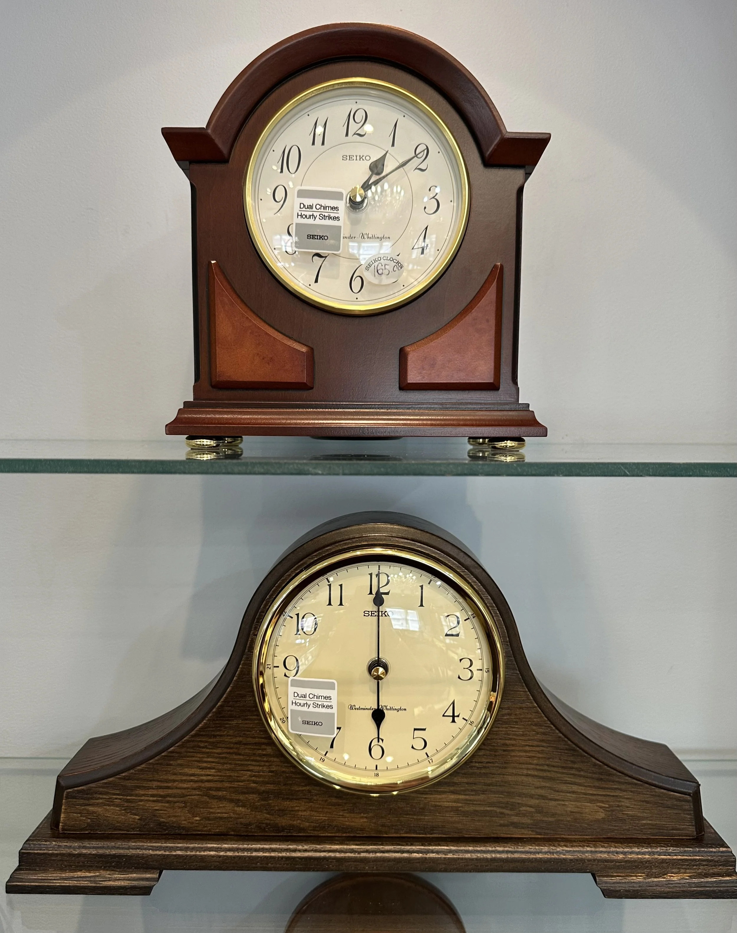 Two vintage-style wooden mantel clocks, one on top of the other, with cream-colored faces and black hands, displayed on glass shelves.