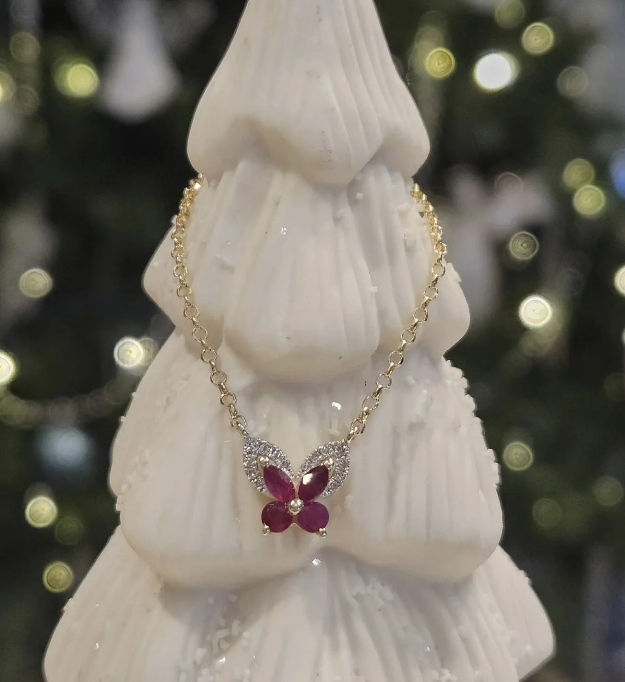 A gold chain necklace with a butterfly-shaped pendant featuring red and pink stones, hanging on a white decorative Christmas tree ornament.