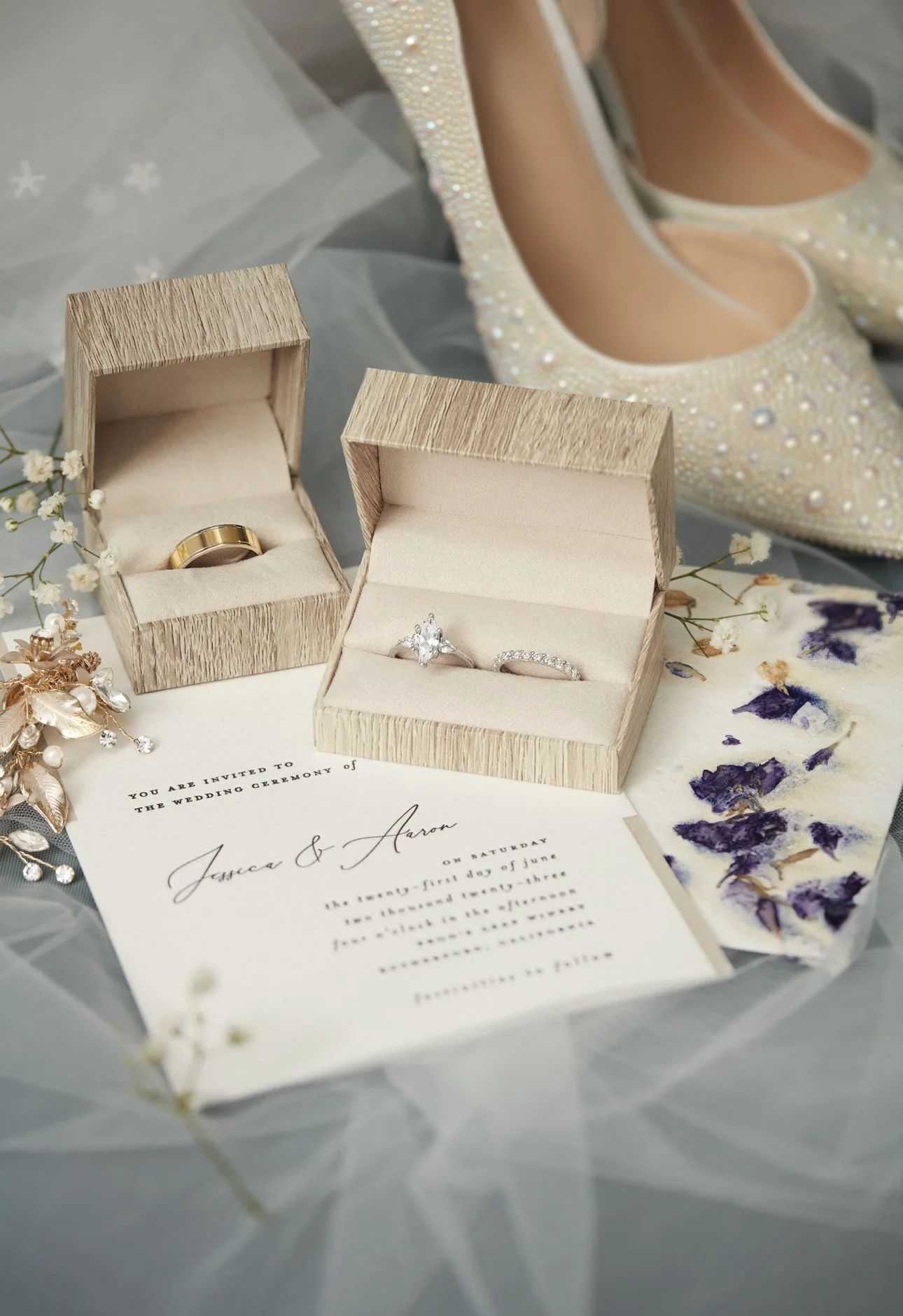 Wedding rings in boxes, a wedding invitation, a pair of embellished wedding shoes, and dried flower petals.