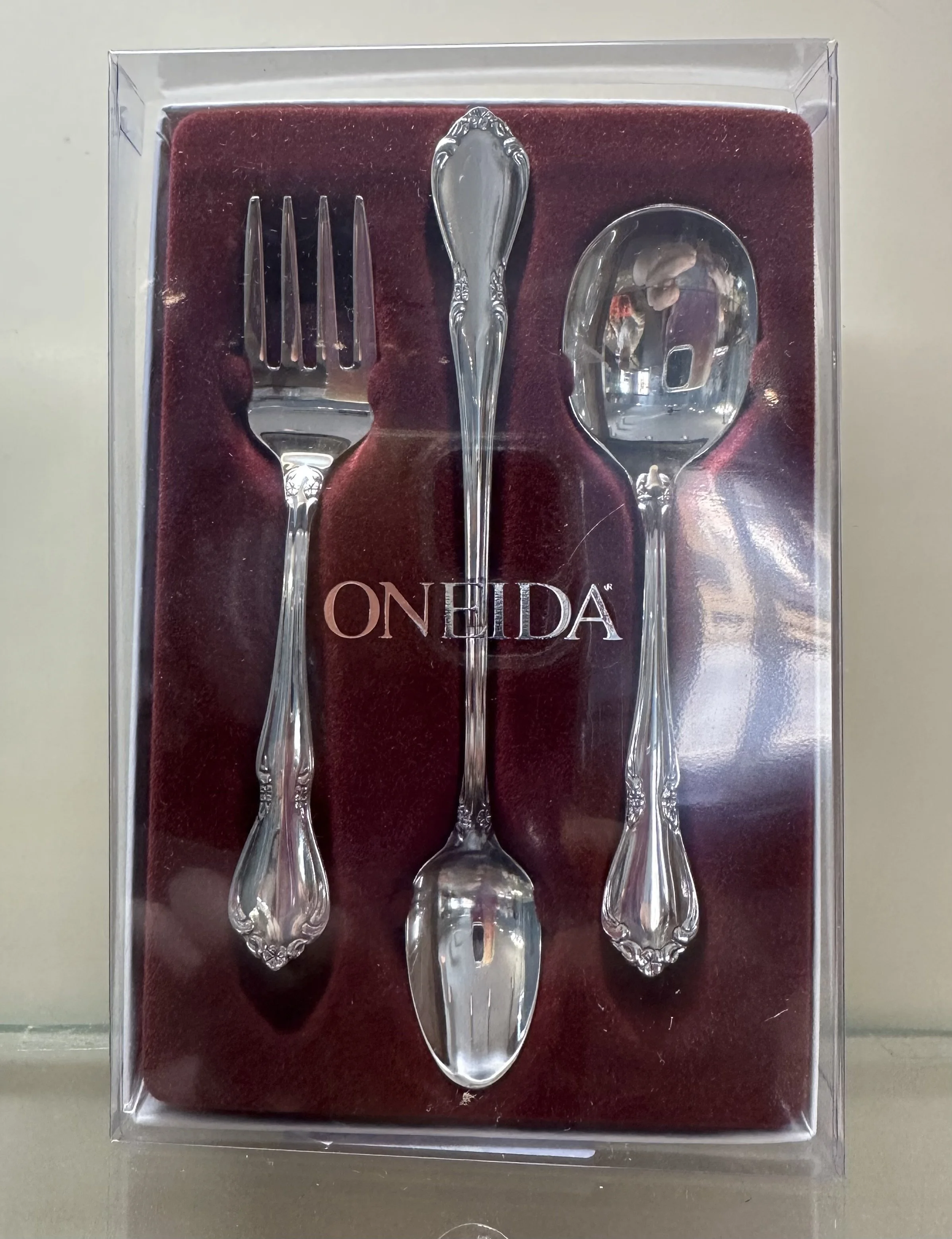 A three-piece silverware set consisting of a fork, a spoon, and a tablespoon with ornate handles, in a plastic display case with a red velvet background and the brand name 'ONEIDA' in the center.