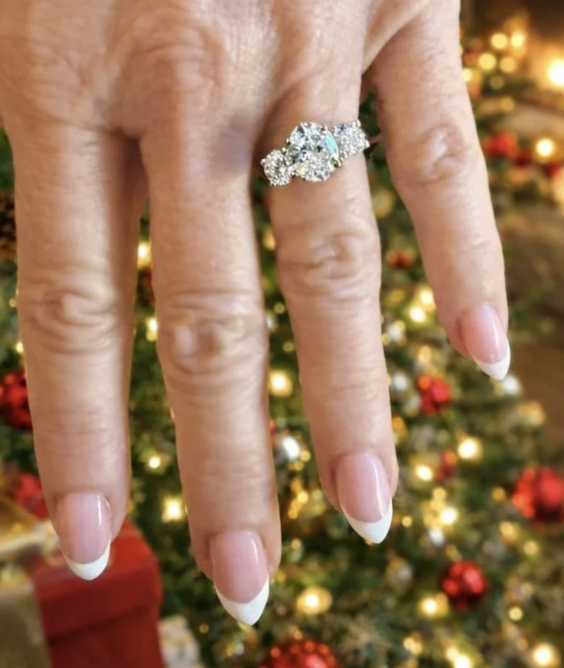Close-up of a hand displaying a sparkling diamond ring on the ring finger, with Christmas tree decorated with lights and ornaments blurred in the background.
