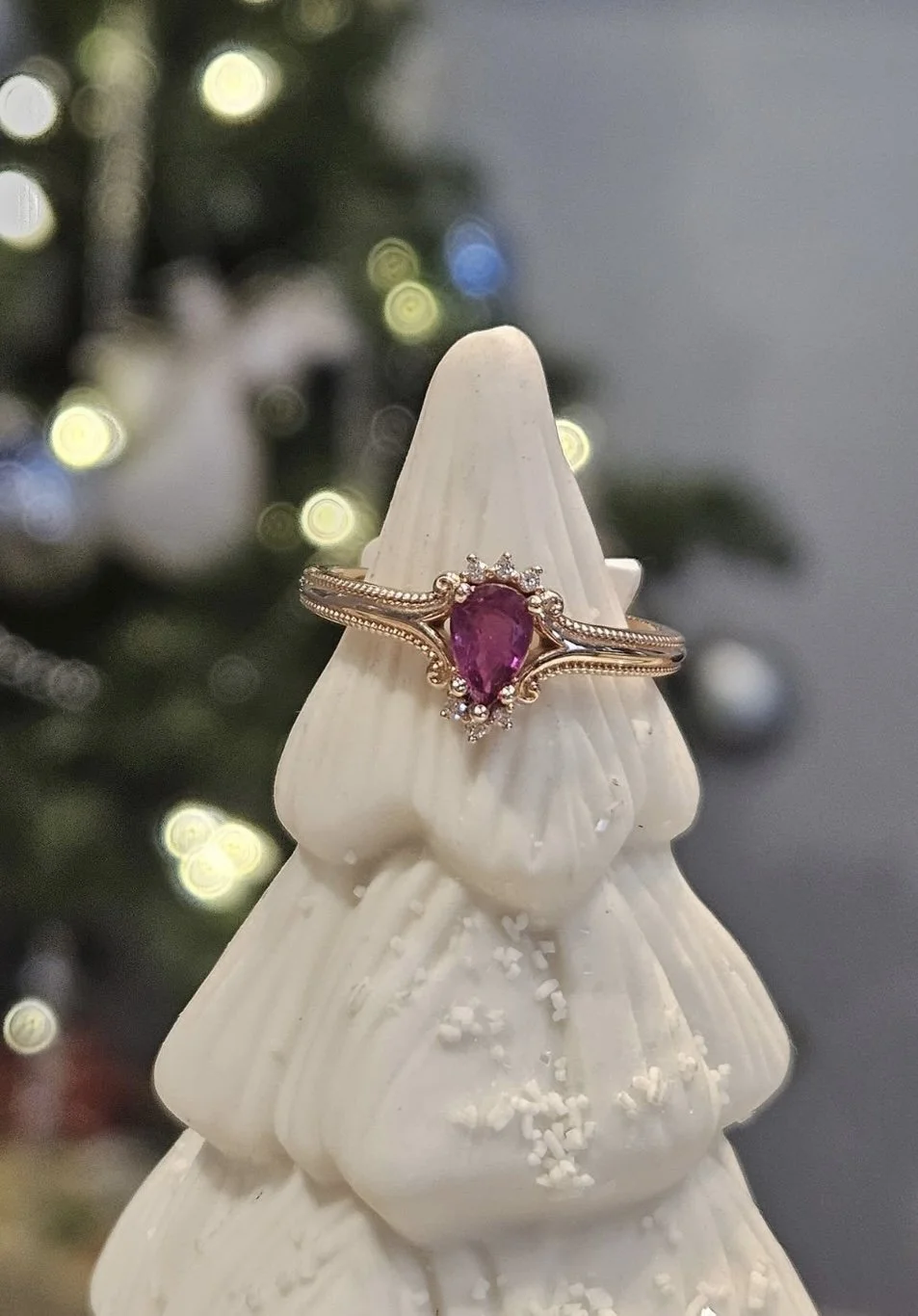 A ring with a teardrop-shaped pink gemstone surrounded by small diamonds, placed on a white ceramic Christmas tree ornament.