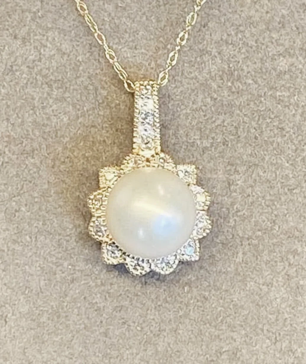 A gold necklace with a large pearl pendant surrounded by small diamonds, hanging on a beige fabric background.
