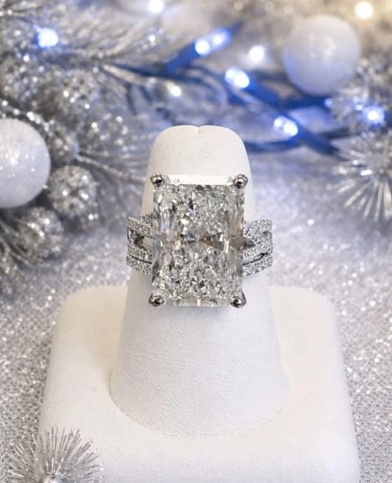 A large, rectangular, diamond engagement ring with a halo setting on a white ring display, surrounded by silver holiday decorations, including glittery ornaments and tinsel.