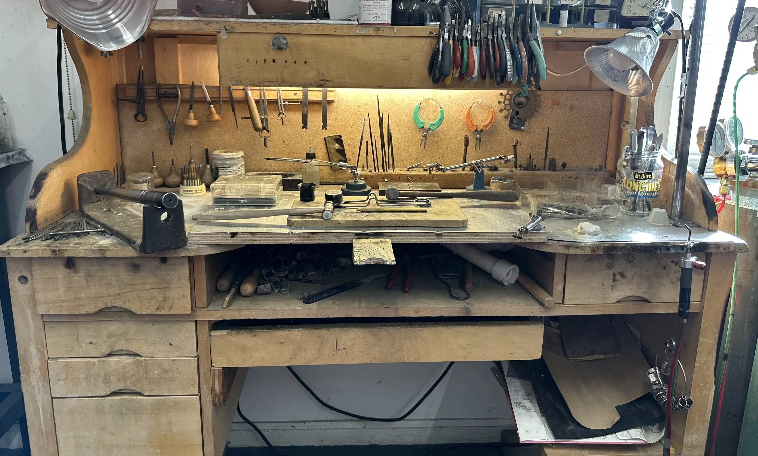 A well-used jewelry workbench with various pliers, files, and small tools hanging on a pegboard. The surface holds small containers, a beading block, and scattered jewelry-making supplies. The back and sides of the workbench are wooden, with drawers and a lower shelf containing additional tools and materials.
