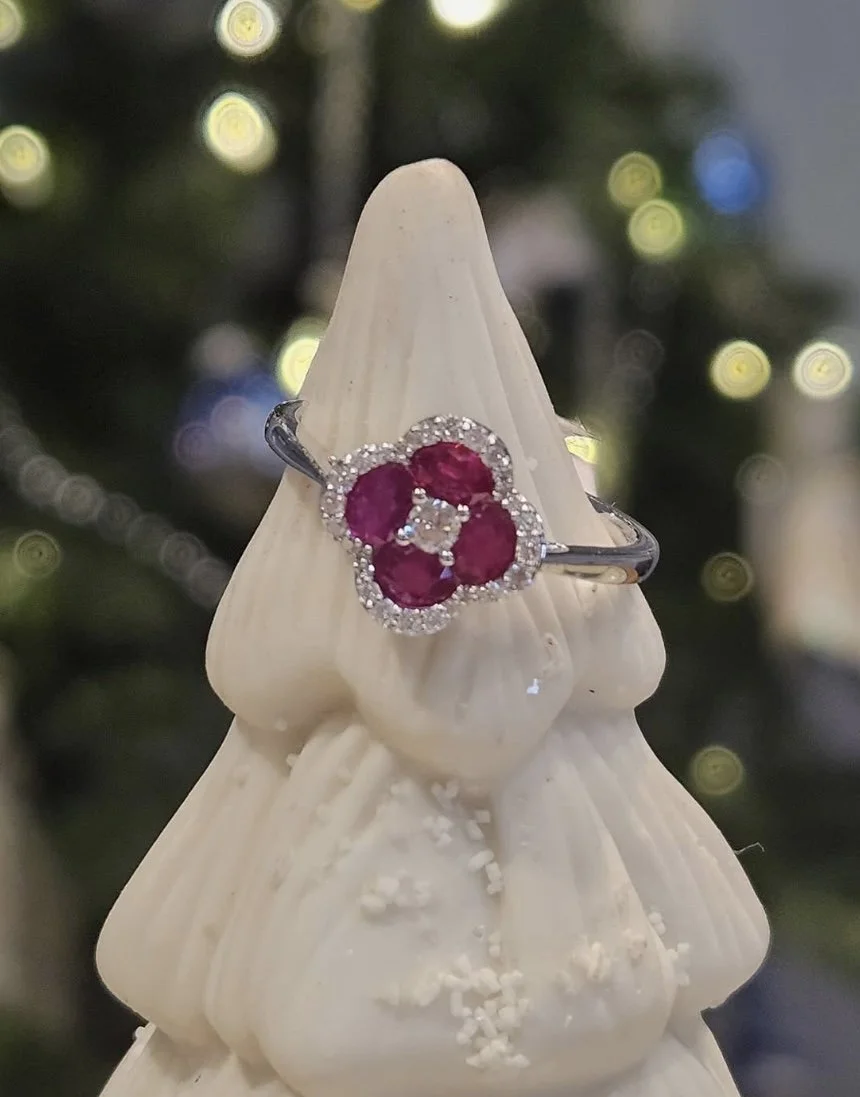 A ring with a flower design of four pink gemstones surrounded by small clear stones, displayed on a cone-shaped white object with a blurred background of Christmas lights.