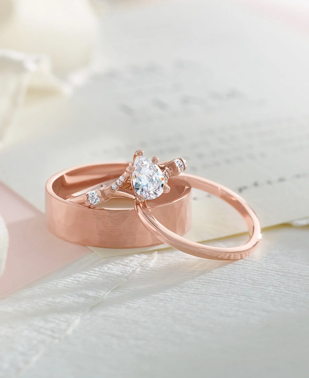 A close-up of a rose gold engagement ring with a large teardrop-shaped diamond, alongside a matching plain band, resting on a satin surface with a blurred background.