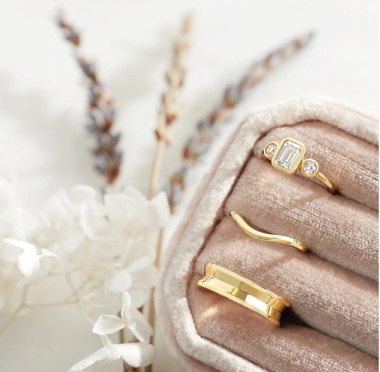 Close-up of a pink velvet jewelry box containing three gold rings with gemstones, surrounded by white flowers and dried lavender.