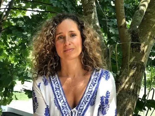 Cara Jordan, a Doula & Night Nanny in West London, Maidenhead & Reading, sits next to the Thames in Maidenhead, smiling and wearing a white blouse with a blue print. There is green foliage behind her.