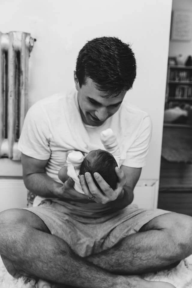 A new father sits cross legged on the floor, cuddling his newborn baby, in W14, London. The man is dressed in shorts and has dark hair.