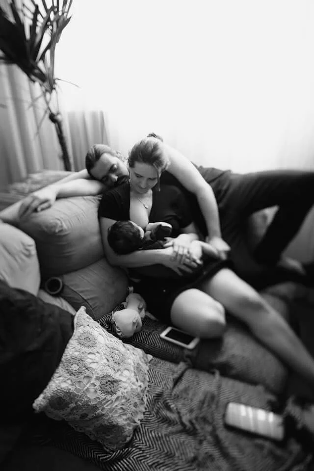 A woman is breastfeeding her baby whilst reclining on a sofa in Kensington, London, her husband sleeps on the sofa behind her.