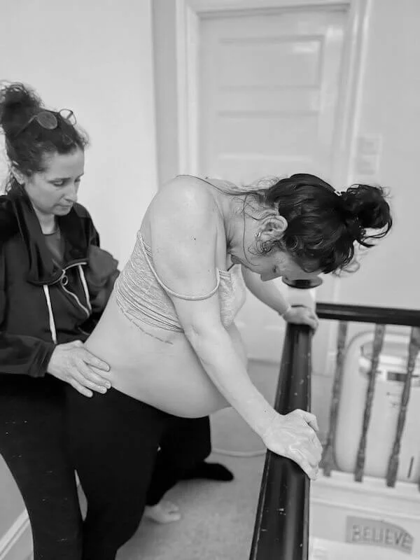 A birth doula, Cara Jordan Birth Doula in London, squats with her client who is in labour, Cara applies counter pressure to the labouring woman's back as a birth comfort technique.