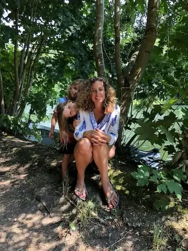 Cara Jordan, a Doula & Night Nanny in West London, Maidenhead & Reading, sits next to the Thames in Maidenhead with her children, all are smiling and Cara is wearing a white blouse with a blue print.