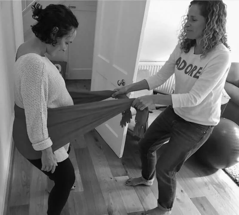 Cara Jordan, a birth doula, demonstrates a pregnancy and birth comfort technique, to her client in Oxford, using a Rebozo Scarf.  Both women have curly hair and are standing, the pregnant woman relaxes with the rocking motion of the scarf.